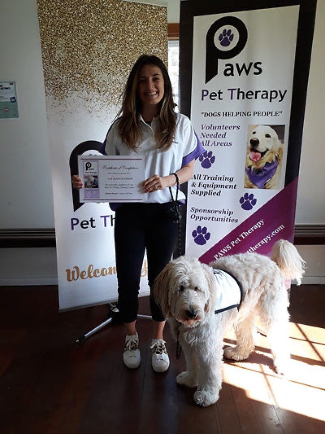 A volunteer with her certificate and dog beside the PAWS banner