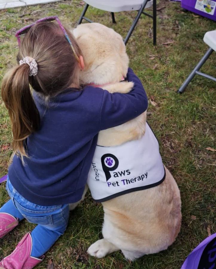 A child hugging a PAWS therapy dog wearing its vest, at an outdoor event