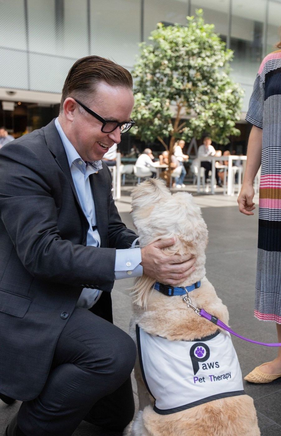 A staff member in a suit greeting an aged care resident and her PAWS dog