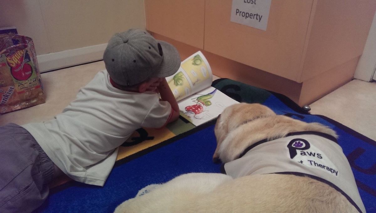 A child reading aloud beside a PAWS therapy dog in a library, part of the PAWS'n'Tales program